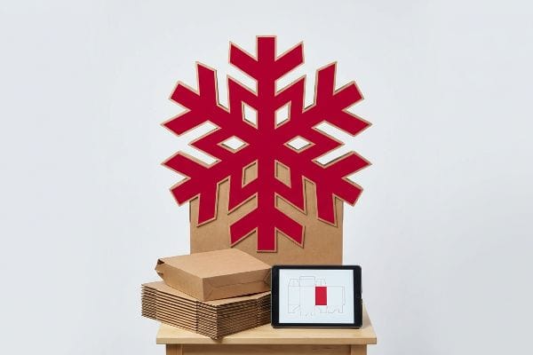 Red die-cut snowflake cardboard display for seasonal product promotion, with flat-packed brown boxes and a tablet showing a packaging design.