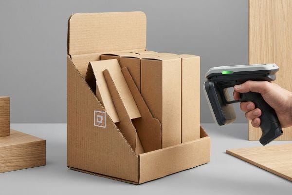 Ein von einer Hand gehaltener RFID-Scanner scannt braune Wellpapp-Displaykartons mit einem Square-Logo, wobei eine grüne Kontrollleuchte aufleuchtet.