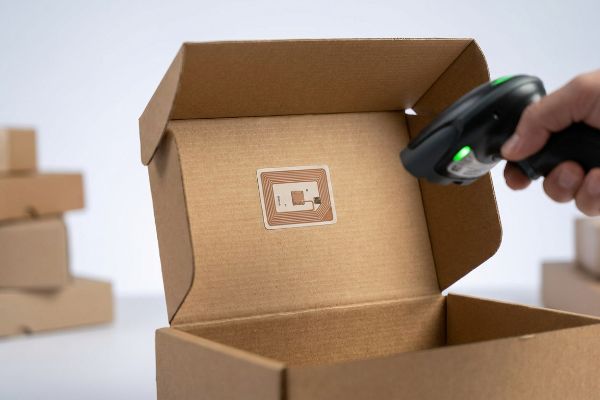 Brown corrugated cardboard box interior with a white RFID tag, scanned by a green-lit handheld device.
