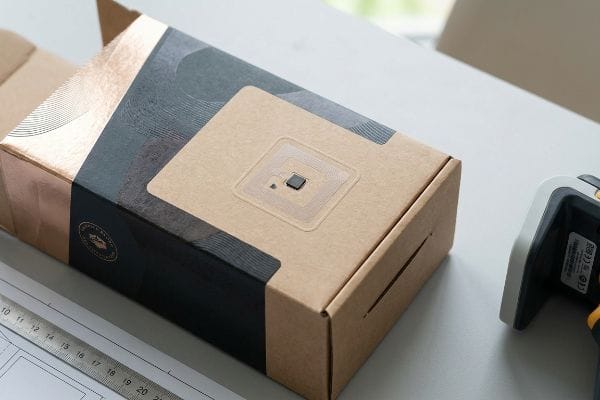 Brauner Kraftkarton mit RFID-Tag, Mikrochip und Antenne auf einer transparenten, metallfreien Zone, neben einer Verpackung aus Metallfolie.