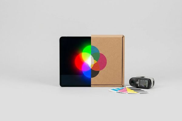 Tablet with RGB color wheel, cardboard box with CMYK color wheel, spectrophotometer, and CMYK swatches for print proofing.