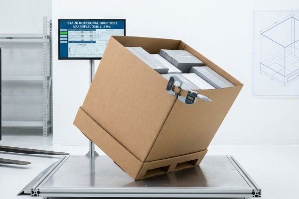 A digital micrometer measures deflection on a tilted brown cardboard box, filled with grey blocks, undergoing an ISTA 2B Rotational Drop Test, with Max Deflection: 21.5 MV displayed.
