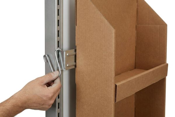 A hand attaches a universal metal S-clip to a gondola bracket, securing a brown corrugated cardboard side cap display.