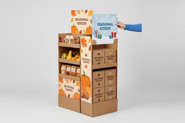Seasonal Display Sign Swap A hand places a winter 'Seasonal Goods' sign on a corrugated cardboard display, transitioning from autumn pumpkins and gourds to holiday boxes.