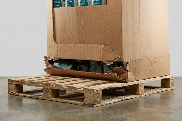 Brown corrugated cardboard display box sagging into a gapped wooden pallet, revealing Luxury Shampoo bottles.