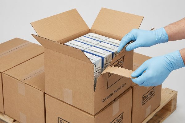 Uma pessoa com luvas azuis abre uma caixa de papelão ondulado, pronta para exposição, utilizando a fita perfurada para rasgar, revelando