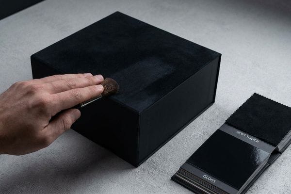Eine Hand streicht über eine schwarze, mit Soft-Touch-Oberfläche beschichtete Schachtel, daneben ein Farbmusterbuch mit Soft-Touch- und Glanz-Oberflächen.