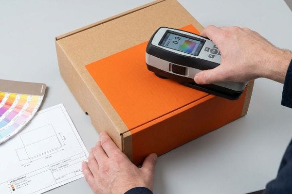 An orange spot color patch on a corrugated box is measured by a spectrophotometer displaying 0.9 Delta E, alongside a Pantone swatch.