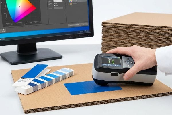 Pantone CMYK Color Measurement A handheld spectrophotometer displays Delta-E 1.8 while measuring a blue Pantone color swatch on corrugated cardboard, ensuring precise CMYK conversion.