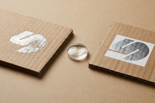 Corrugated fiberboard pieces display metallic silver logos, one with a primer base, alongside a clear optical lens.