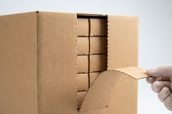 A gloved hand tears open a brown corrugated shelf-ready packaging box, revealing smaller inner cardboard boxes stacked inside.