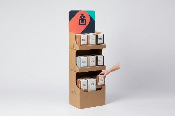 Brown corrugated cardboard three-tier merchandiser with die-cut header. Hand reaches for Premium Coffee box.