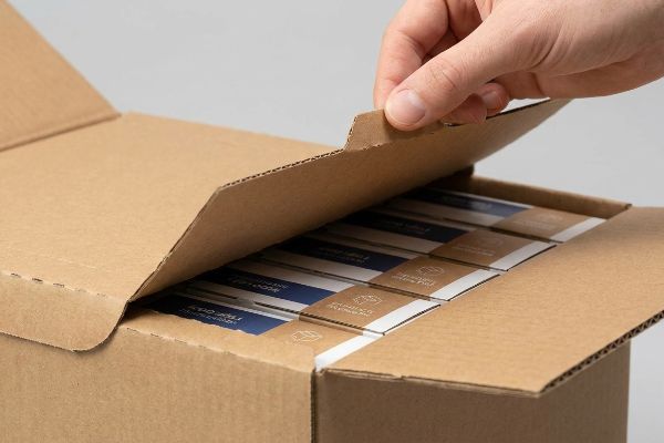 Hand opening a brown corrugated shelf-ready box, revealing multiple product boxes with blue and kraft designs inside.
