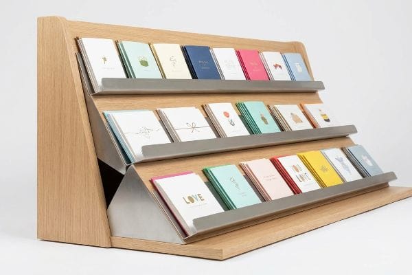 Light oak wood tiered display stand with three angled metallic shelves, preventing greeting card slump and maximizing visibility.