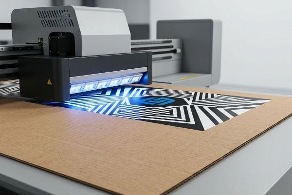 Corrugated cardboard sheet being printed with a black, white, and blue geometric pattern by a UV flatbed printer, showcasing instant ink polymerization.