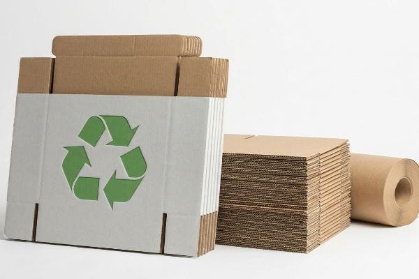 Recyclable corrugated cardboard packaging featuring a green recycling symbol on a white box, alongside stacks of flat brown cardboard sheets and a roll of kraft paper.