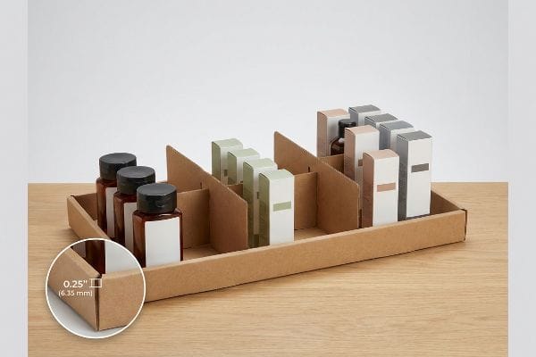 Brown corrugated cardboard display tray with modular dividers separating amber bottles and various product boxes, featuring a 0.25-inch (6.35 mm) clearance.