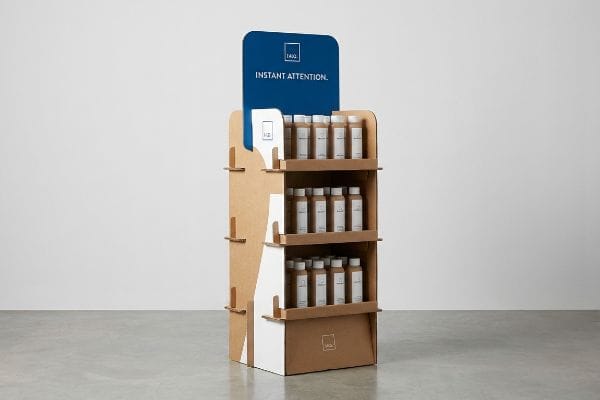 Corrugated cardboard POP display with a blue '1402 Instant Attention' header, showcasing three shelves of small brown product bottles.