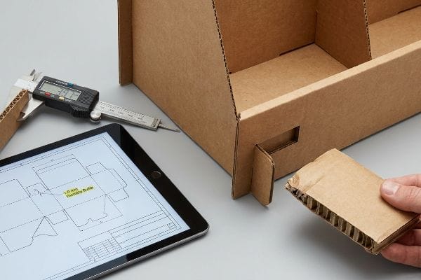 Digital caliper measures corrugated board at 3.97 mm, next to a tablet showing a dieline with a 1.0 mm Humidity Buffer.