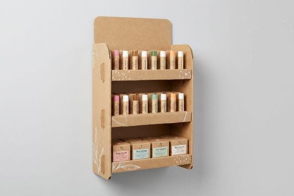 A corrugated cardboard sidekick display showcases lip balms, lip scrubs, lip masks, and lip oils, featuring white leaf patterns.