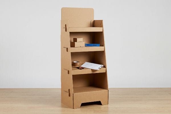 Corrugated cardboard point-of-purchase display with four shelves, holding small kraft boxes, tape, and a clipboard on a wooden floor.