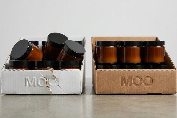 Debossed Tray Integrity Debossed Moo logo on two corrugated trays, one white damaged, one kraft intact, holding amber glass jars with black lids.