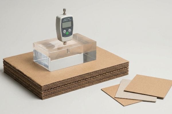 Corrugated Board Load Test A digital force gauge performs a Max Load Capacity Test on a stack of fluted corrugated board, with individual cardboard samples nearby.
