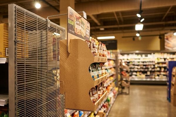A brown corrugated power wing display, filled with snack bags, hangs from a metal wire rack in a grocery store aisle, showcasing retail merchandising.