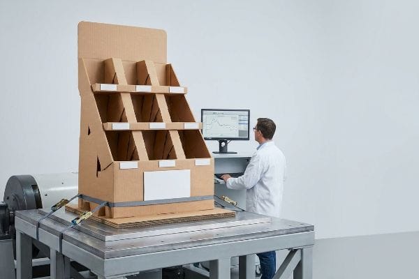 Display Vibration Testing Brown corrugated cardboard display undergoing ISTA 3A dynamic vibration testing on a kinetic table, secured with straps, as a technician monitors structural data on a computer.