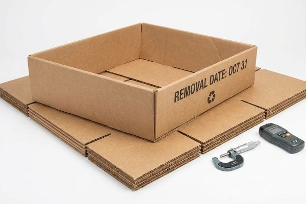 Corrugated cardboard display box with "Removal Date: Oct 31" and recycling symbol, next to stacked flat pieces and a micrometer.