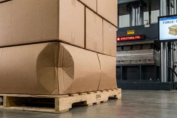 パレットの張り出し部分の構造的欠陥 GMAパレットに積まれた段ボール箱は、油圧圧縮試験中に「構造的欠陥」を示し、一番下の箱が突き出た部分から明らかにたわんでいる。.