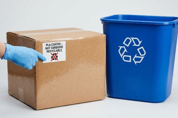 PLA Coated Not Recyclable Hand in blue glove points to 'Pla Coated - Not Curbside Recyclable' label on a cardboard box next to a blue recycling bin.
