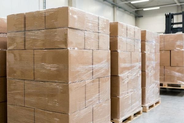 Warehouse with multiple stacks of shrink-wrapped brown cardboard boxes on wooden pallets, forklift visible.