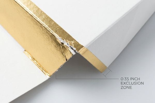 منطقة نفخ النقش ورق ذهبي على زاوية علبة مستحضرات تجميل بيضاء بها تمزق، مما يسلط الضوء على منطقة الاستبعاد 0.35 بوصة.