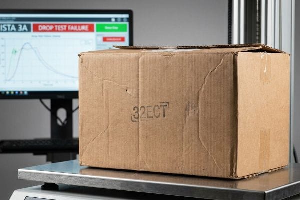 Box Drop Test Failure Brown corrugated cardboard box with a '32ECT' stamp on a digital scale, in front of a monitor showing 'Ista 3a' and a 'Drop Test Failure' alert.