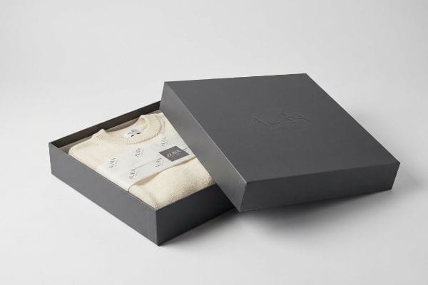 A dark grey premium gift box, partially open, reveals a folded cream Aura sweater with a branded paper band and embossed Aura logo on the lid.