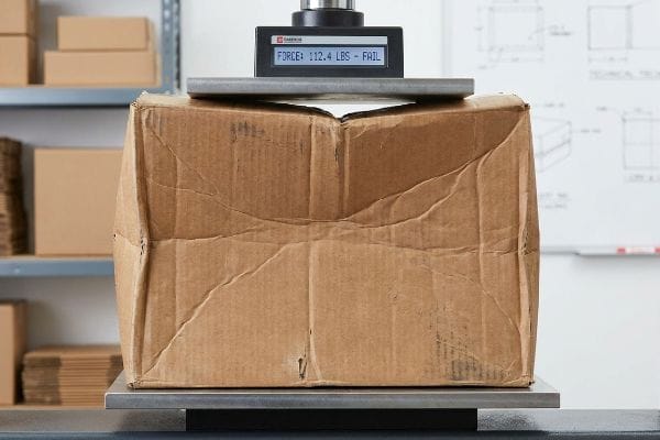 Mailer Fatigue Failure Test Crumpled brown corrugated box failing a compression test, displaying 'Force: 112.4 Lbs - Fail' on the machine, highlighting fiber fatigue.