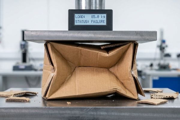 Packaging Failure Test Corrugated cardboard box failing a compression test under a hydraulic press, displaying 'Load: 85.0 kg Status: Failure' on the digital screen.
