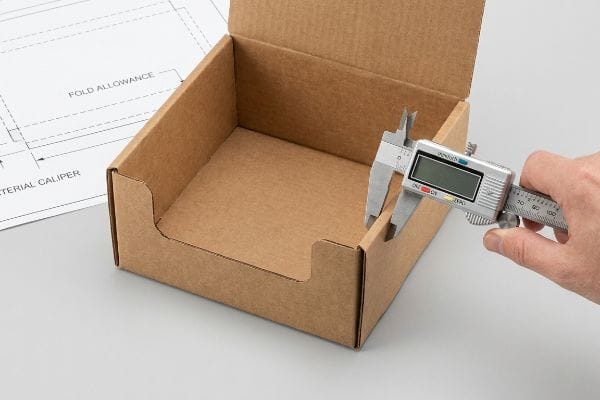 Measuring a brown corrugated cardboard box's material thickness with a digital caliper, alongside a technical drawing for 'Fold Allowance' and 'Material Caliper'.