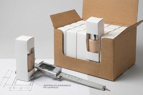 Digital caliper measures a white SBS carton with foundation bottle, showing 'Micro-Clearance Required' on a blueprint, next to a retail tray.