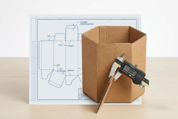 A digital caliper measures 3.00mm corrugated cardboard next to a hexagonal box and its blueprint showing Caliper Compensation and 120-degree angles.