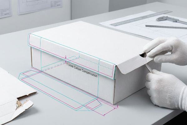 Gloved hands assemble a white corrugated box with cyan and magenta design overlays, highlighting 1.5mm Caliper Compensation for precise folding.