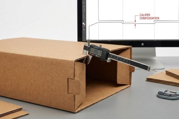 Brown corrugated cardboard box measured by a digital caliper, with a monitor displaying a dieline illustrating Caliper Compensation for precise fold allowances.