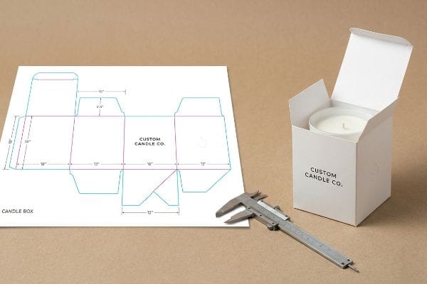 Candle Box Dieline Design A white paper displays a detailed Candle Box dieline with structural geometry and measurements, alongside an open Custom Candle Co. candle box and a silver caliper for precise packaging design.