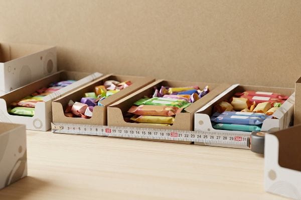 Colorful wrapped candies in brown cardboard display boxes are measured by a white tape, showing retail countertop tray sizes.