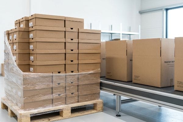 Warehouse scene with cardboard shoe boxes on a shrink-wrapped pallet and Master Shipper boxes on a conveyor.