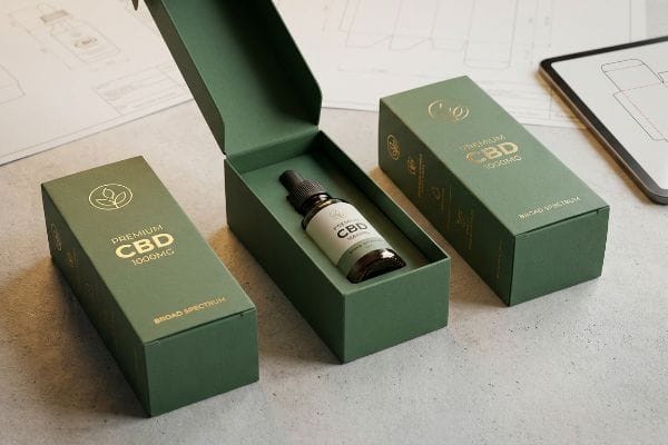 Maquete de embalagem de caixa de CBD Caixas personalizadas verdes de CBD Premium de 1000mg de amplo espectro com logotipos em folha de ouro, uma delas aberta revelando um frasco conta-gotas, sobre uma mesa com plantas de design.