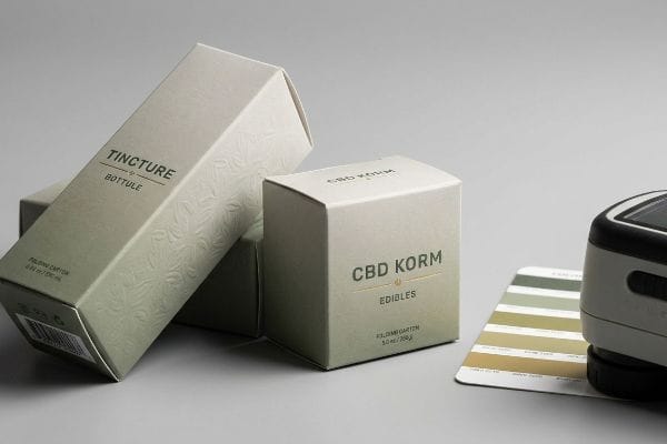 CBD Packaging Color Measurement Embossed folding cartons for Tincture Bottule and CBD Korm Edibles, with a spectrophotometer measuring color on a swatch card.