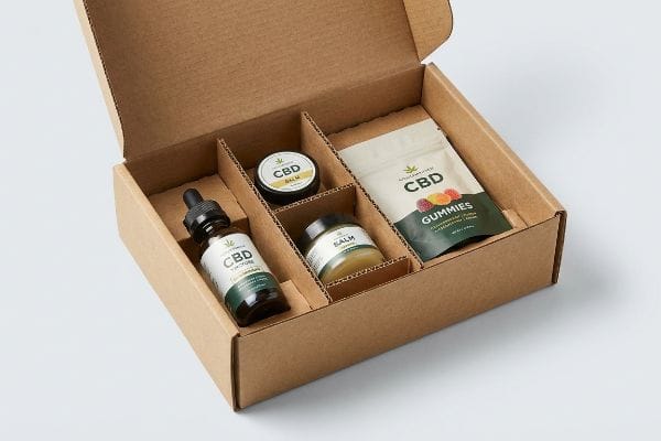Cbd Product Assortment An open corrugated cardboard gift box features modular dividers securing a Cbd Tincture bottle, Cbd Balm, Salm jar, and a pouch of Cbd Gummies.