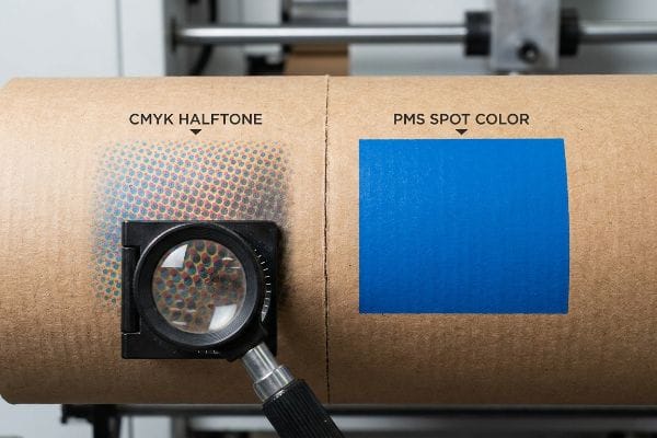 CMYK PMS Print Comparison A loupe magnifier reveals CMYK Halftone dots on corrugated material next to a solid PMS Spot Color blue block, demonstrating print differences.